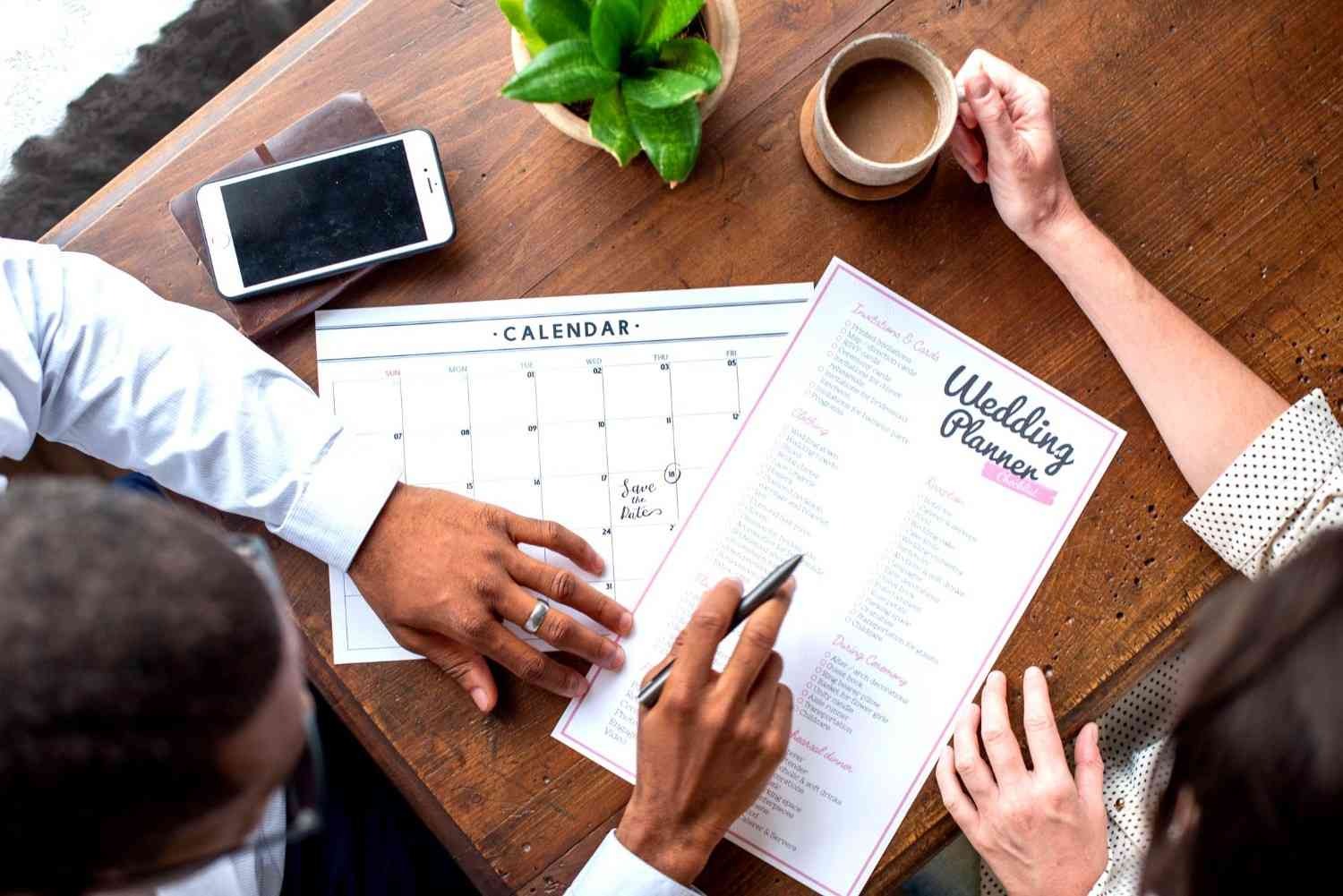 Couple discussing wedding planning checklist and event schedule on a wooden table with calendar and coffee.