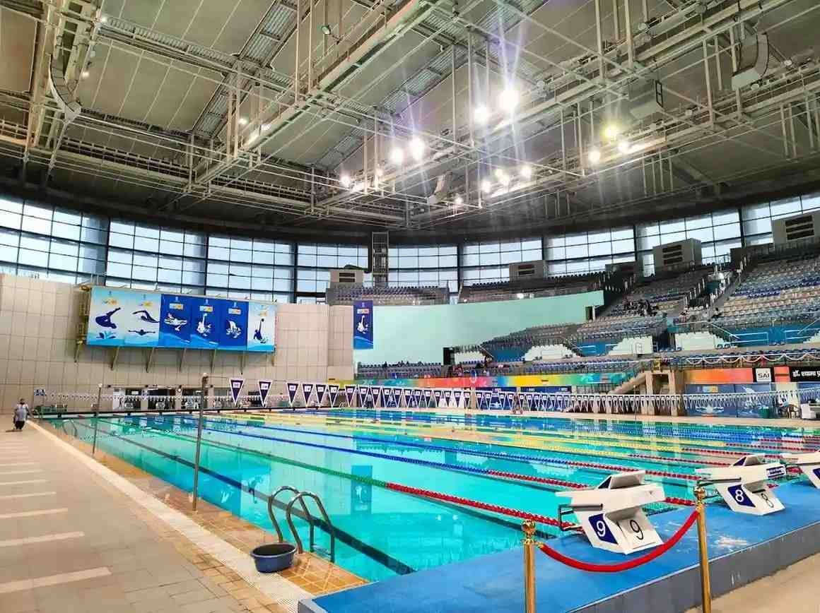 A wide-angle interior view of the Dr. Shyama Prasad Mukherjee Swimming Pool Complex shows an Olympic-sized turquoise pool with multiple lanes.