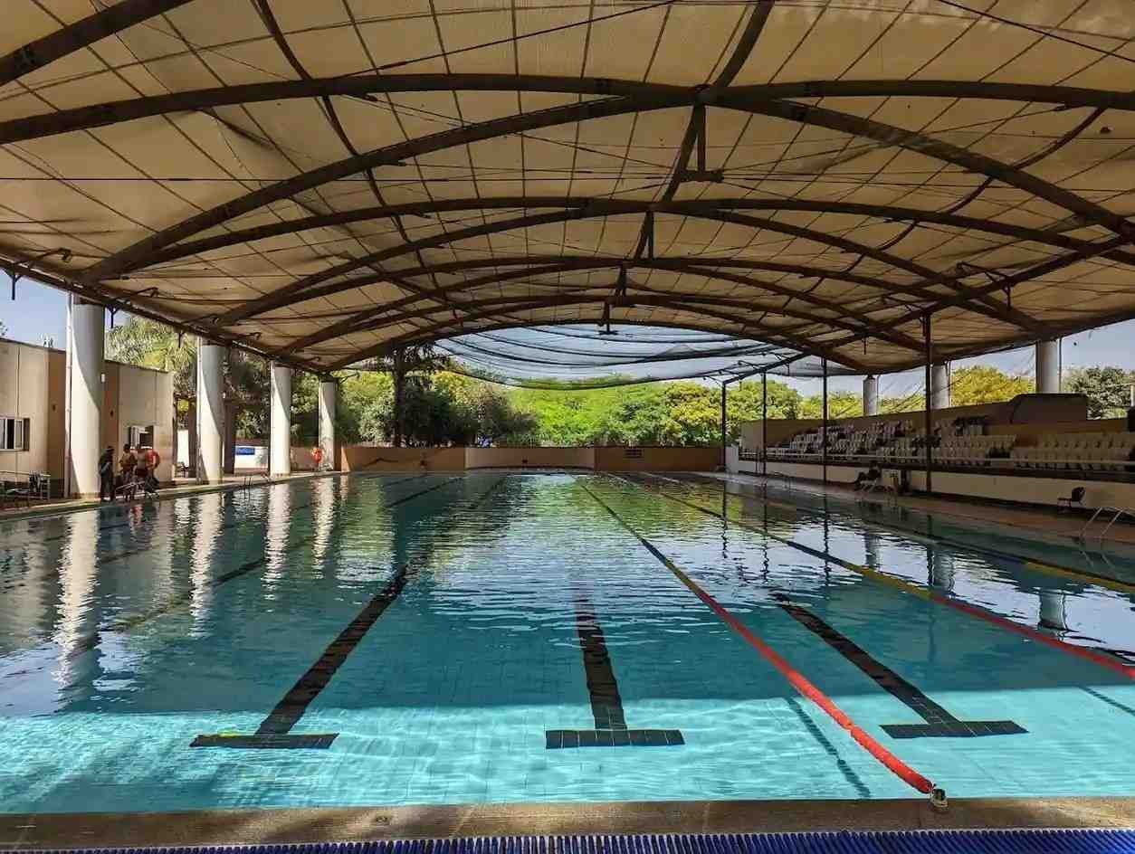 Siri Fort Sports Complex Swimming Pool in New Delhi India: semi-open-air Olympic-sized lap pool with 8 lanes, black lane dividers, clear turquoise water under beige fabric canopy roof with steel arches.
