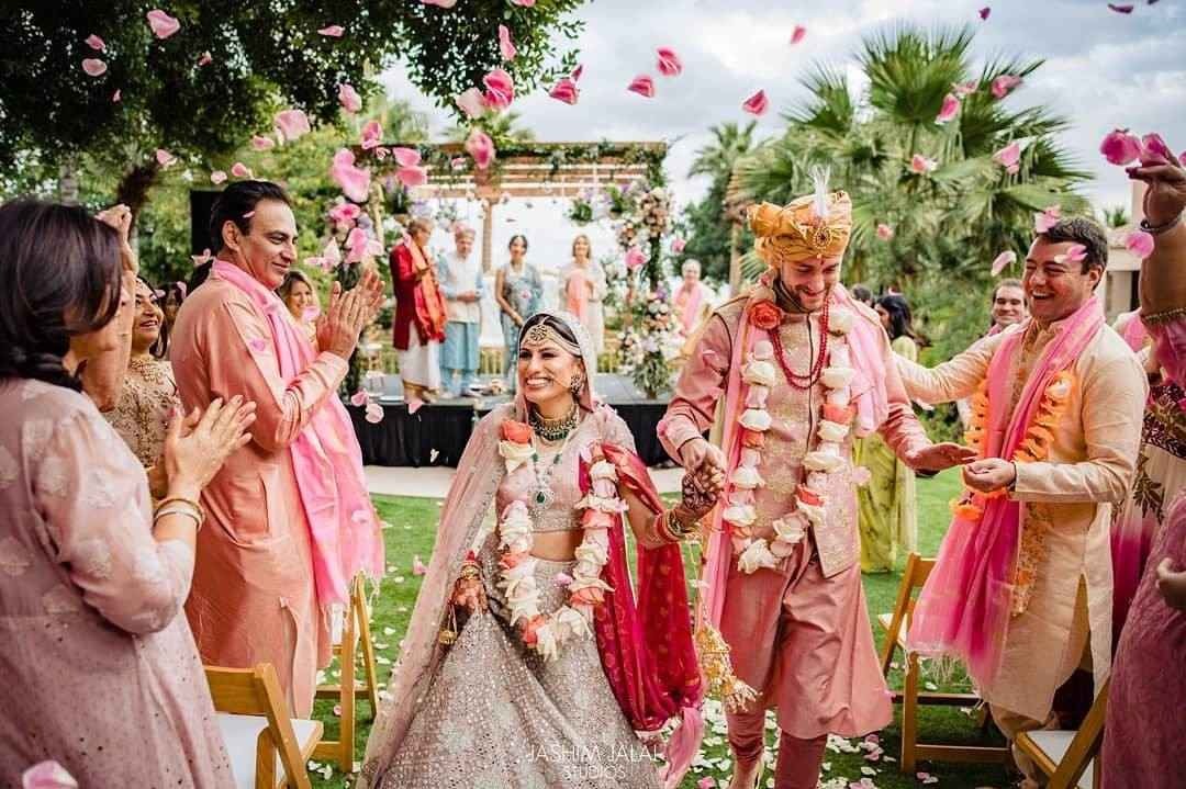 A smiling bride and groom in traditional South Asian wedding attire walk down the aisle as guests cheer and toss pink rose petals during an outdoor ceremony.