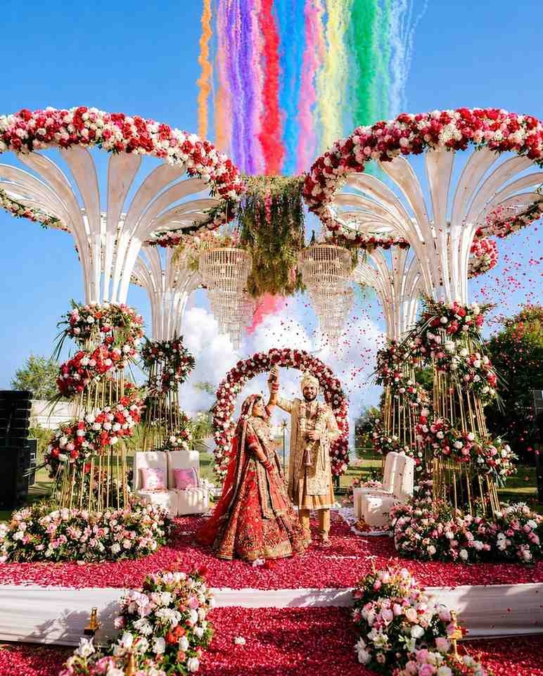 Bride and groom celebrating their wedding on a luxurious outdoor floral stage with colorful confetti, elegant décor, and grand wedding mandap.