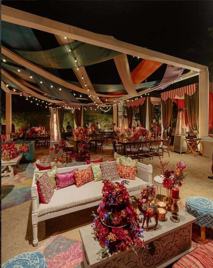 Elegant outdoor wedding venue with colorful draped canopy, floral centerpieces, cozy lounge seating, and warm string lights creating a luxurious evening celebration ambiance.